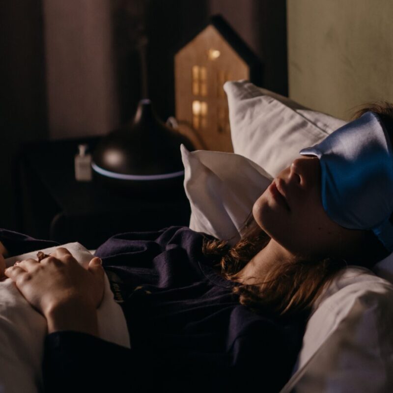 A woman lying in bed wearing a blue sleep mask, peacefully resting with her hands on a blanket, beside a diffuser emitting mist—perfectly illustrating the calming effects of sleep meditation.