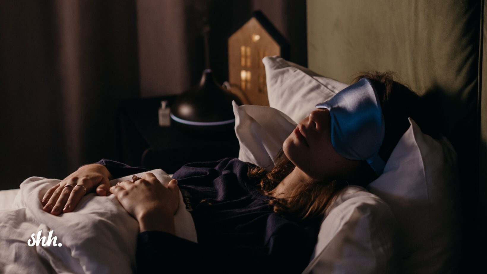 A woman lying in bed wearing a blue sleep mask, peacefully resting with her hands on a blanket, beside a diffuser emitting mist—perfectly illustrating the calming effects of sleep meditation.