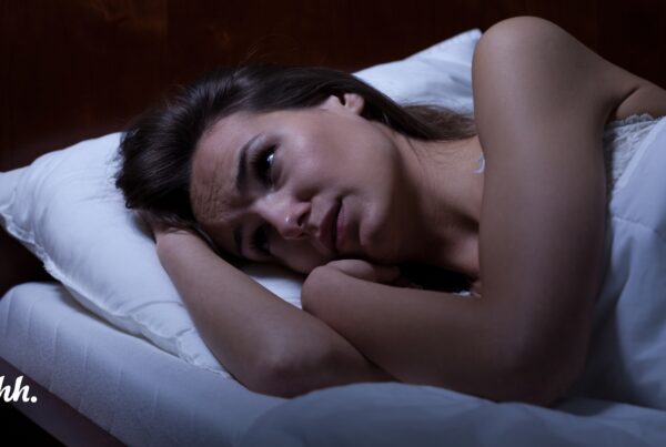 An anxious mom lies awake in bed at night, her eyes open and mind racing with overthinking as she struggles to sleep. The emotional weight of her worries is palpable, yet there's a gentle sense of hope suggesting that a guided meditation could help calm her restless mind.