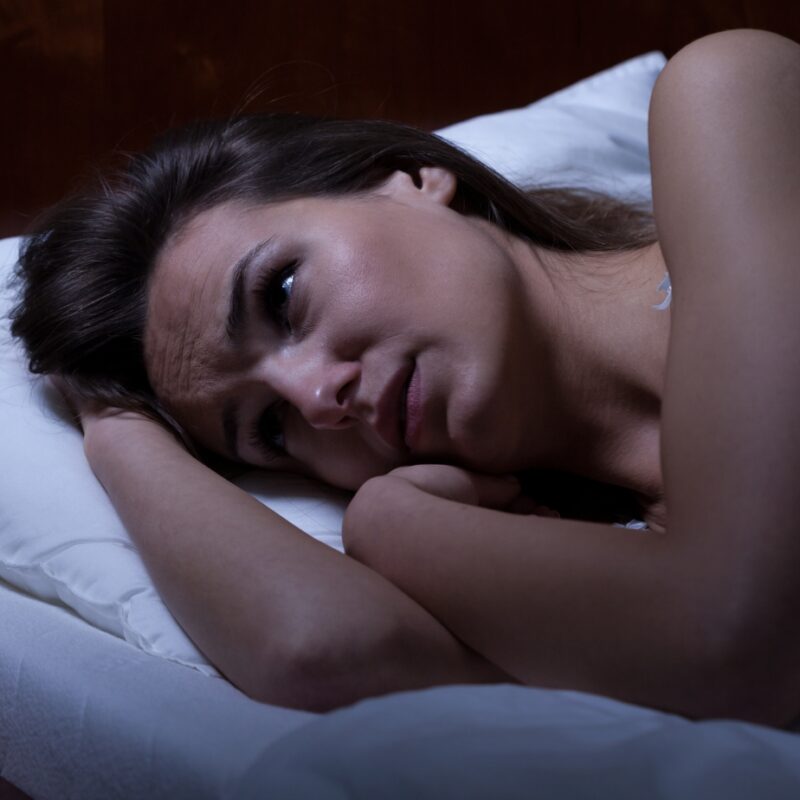 An anxious mom lies awake in bed at night, her eyes open and mind racing with overthinking as she struggles to sleep. The emotional weight of her worries is palpable, yet there's a gentle sense of hope suggesting that a guided meditation could help calm her restless mind.