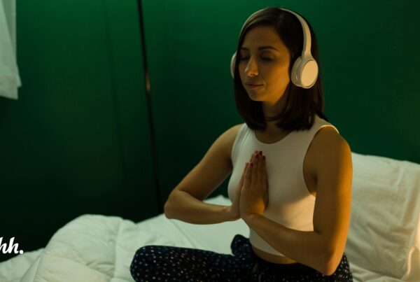 Woman sitting cross-legged on a bed with headphones on, meditating with eyes closed and hands in prayer position—practicing mindfulness with sleep music for relaxation and better rest.