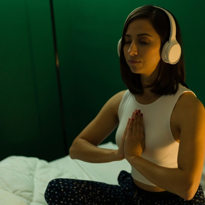 Woman sitting cross-legged on a bed with headphones on, meditating with eyes closed and hands in prayer position—practicing mindfulness with sleep music for relaxation and better rest.