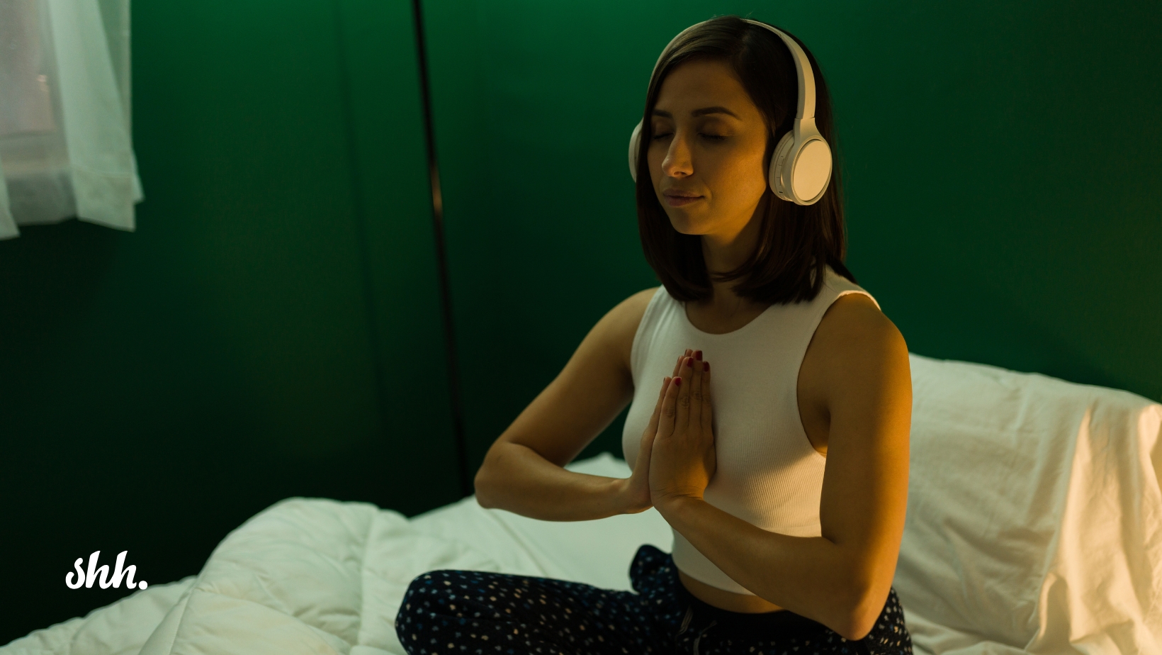 Woman sitting cross-legged on a bed with headphones on, meditating with eyes closed and hands in prayer position—practicing mindfulness with sleep music for relaxation and better rest.
